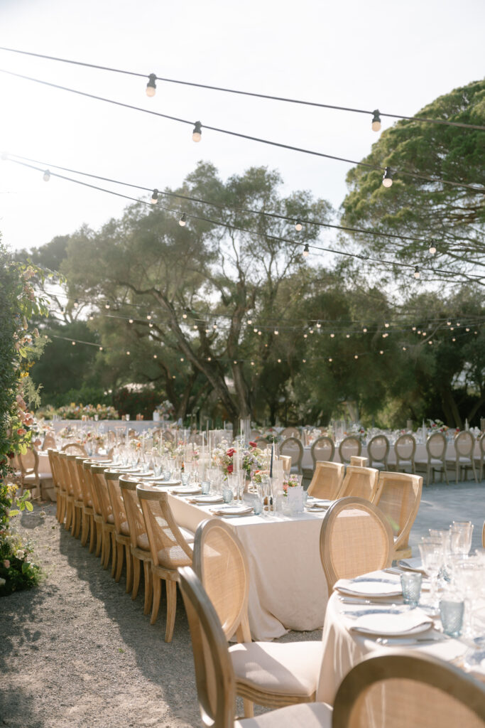 Long banquet tables with beige tablecloths and elegant place settings are arranged outdoors under string lights, surrounded by wooden chairs. Trees and greenery provide a natural backdrop in the sunlit setting.