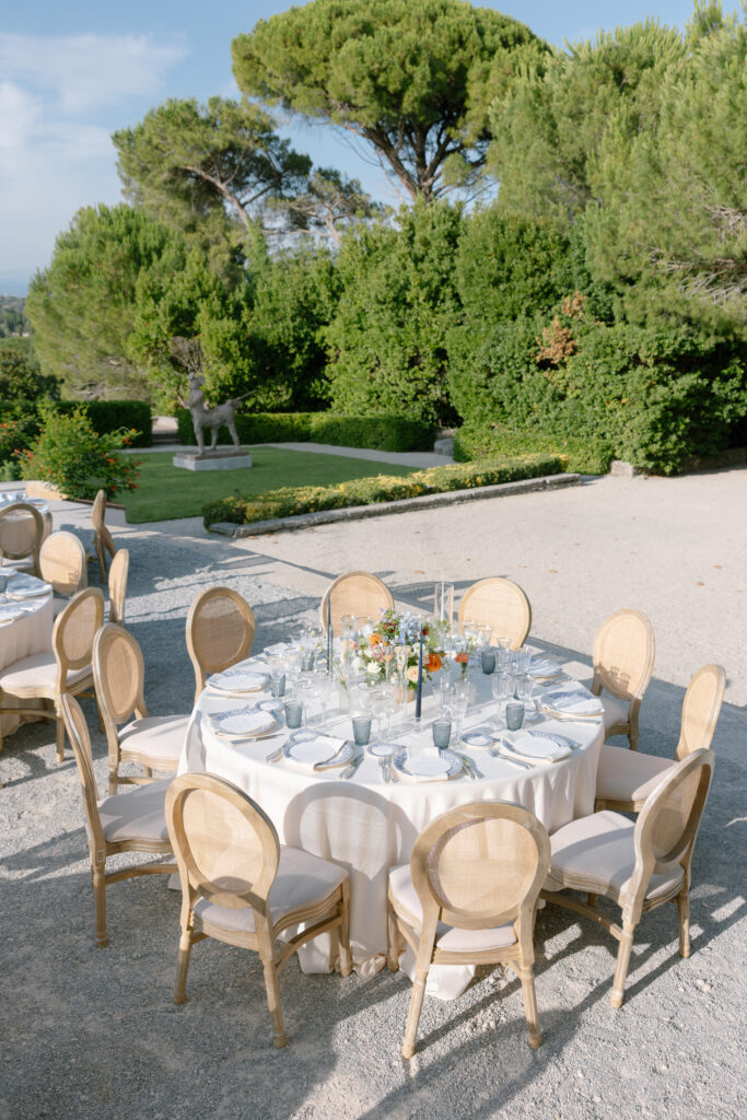 A round table with a white tablecloth and elegant place settings is surrounded by wooden chairs outdoors, with floral centerpieces and lush green trees in the background.