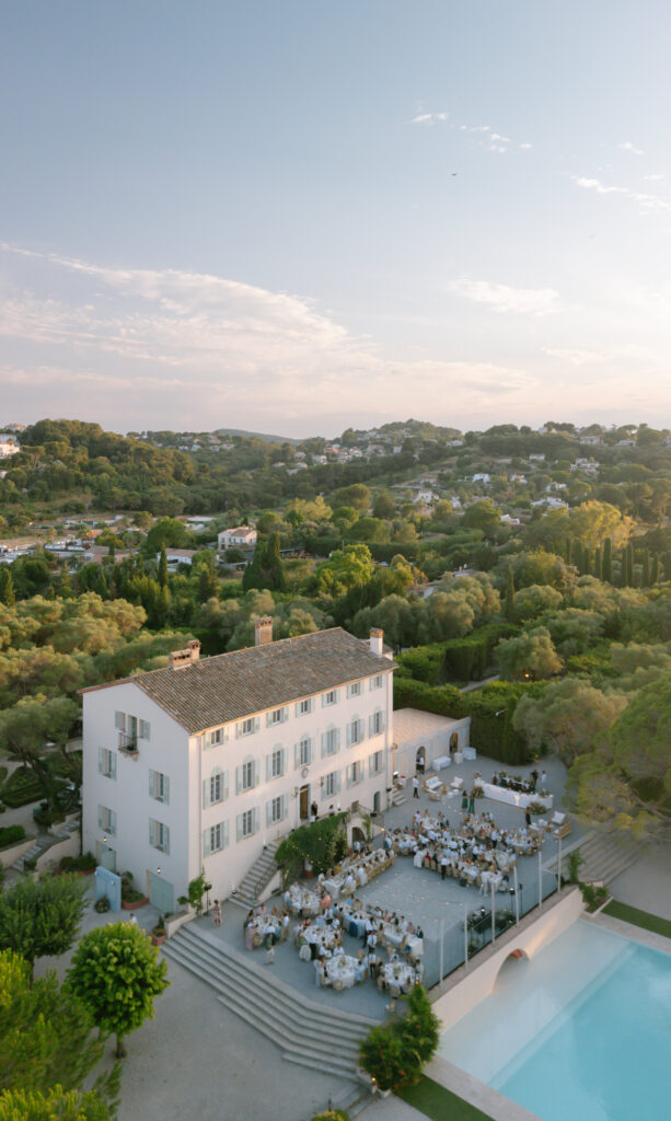 A large white villa with outdoor dining tables set up for an event, surrounded by greenery and hills under a partly cloudy sky, with a swimming pool in the foreground.
