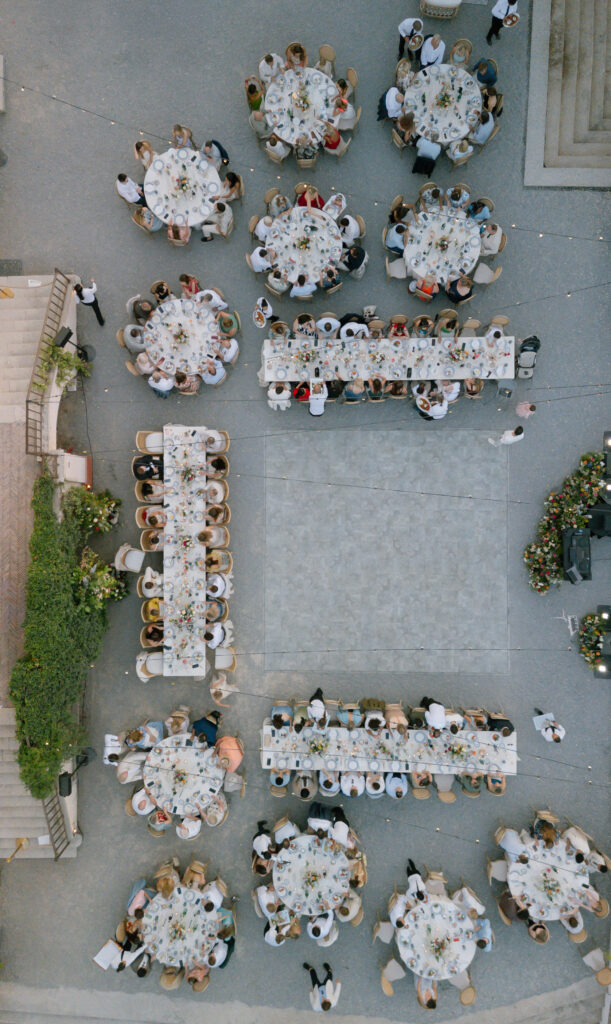 Aerial view of an outdoor event with round and rectangular tables arranged around an open square, surrounded by people dining and mingling. The setting appears elegant and festive.