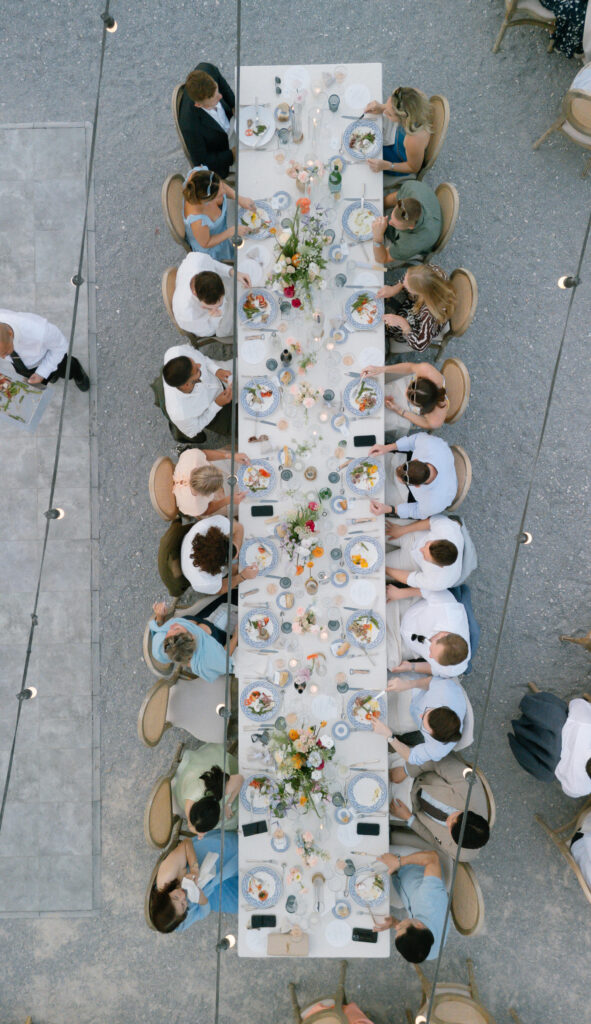 Overhead view of a long outdoor dining table set for a formal meal, with people seated on both sides. The table is decorated with flowers, candles, and plates of food. Warm outdoor lighting and gravel ground are visible.