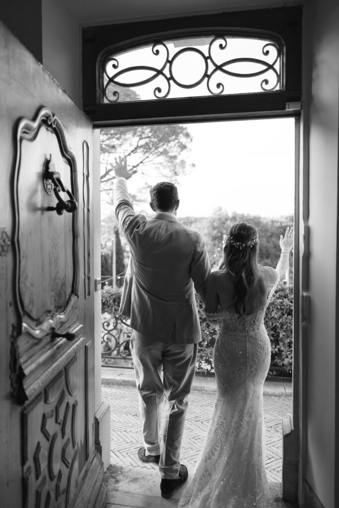 A bride and groom, seen from behind, walk arm in arm through an open ornate doorway, waving as they step outside. The scene is in black and white, capturing a joyful moment after their wedding ceremony.