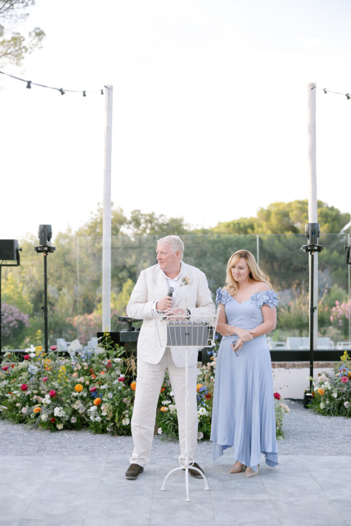 An older man in a white suit and a woman in a light blue dress stand outdoors beside a white metal stand, with flower arrangements and greenery in the background under a clear sky.