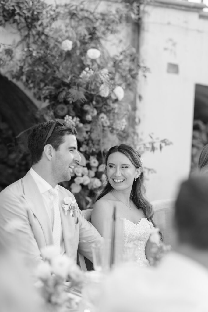 A bride and groom sit side by side, smiling at each other during their wedding reception. The bride wears a strapless dress and the groom is in a light suit, with flowers and greenery in the background. The image is in black and white.