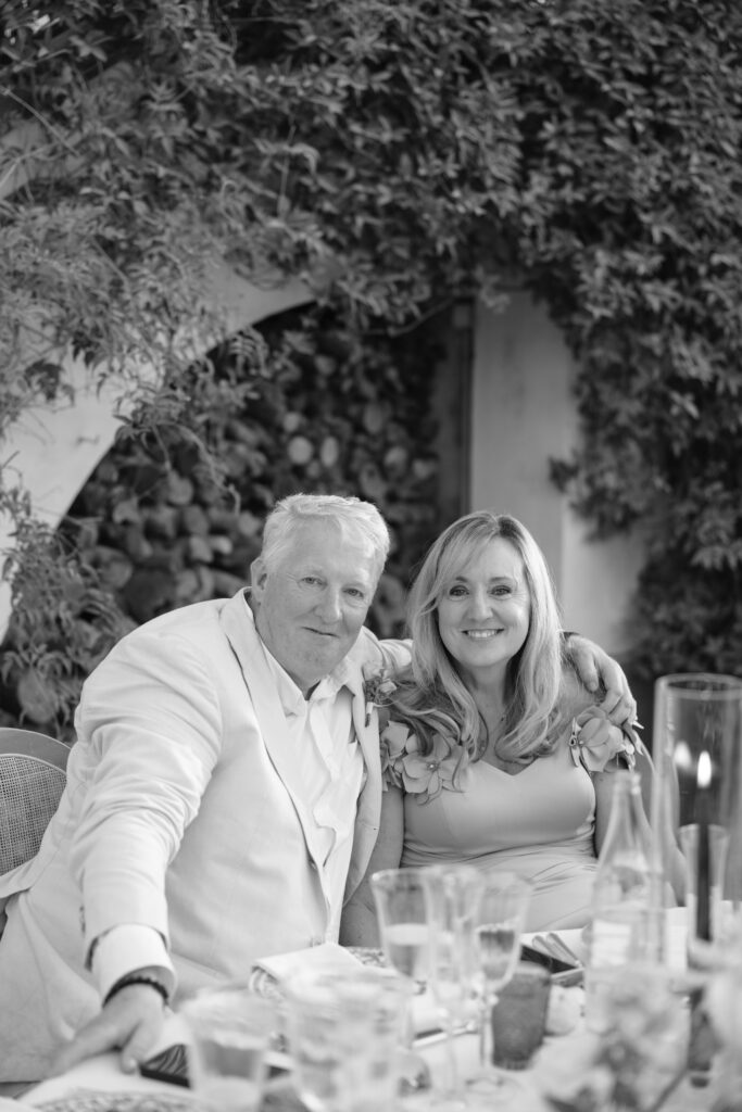 A man and woman sit closely together at a table outdoors, smiling at the camera. The man wears a light-colored jacket; the woman has long, blonde hair and a light dress. Greenery and a stone wall are in the background.
