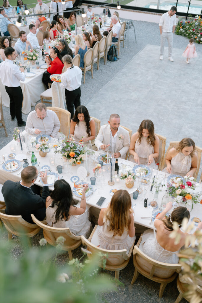 A large group of people dressed in semi-formal attire gather around elegantly decorated tables at an outdoor event, likely a wedding reception, with flowers, candles, and food on the tables.