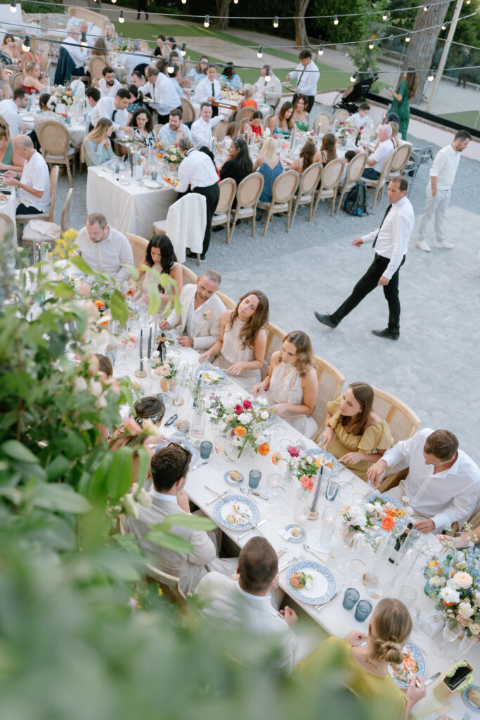 Aerial view of guests seated at long, decorated tables during an outdoor wedding reception, with flowers, candles, and plates of food; a waiter walks nearby under string lights.