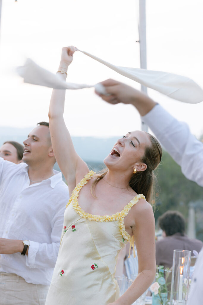 A woman in a light-colored dress joyfully waves a napkin in the air among a group of people at an outdoor event, smiling and celebrating. Others nearby also wave napkins, and candles decorate the table.