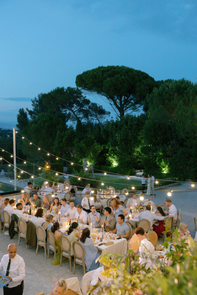 A large group of people dine at long tables outdoors in the evening, surrounded by greenery and trees, with string lights overhead creating a warm and festive atmosphere.
