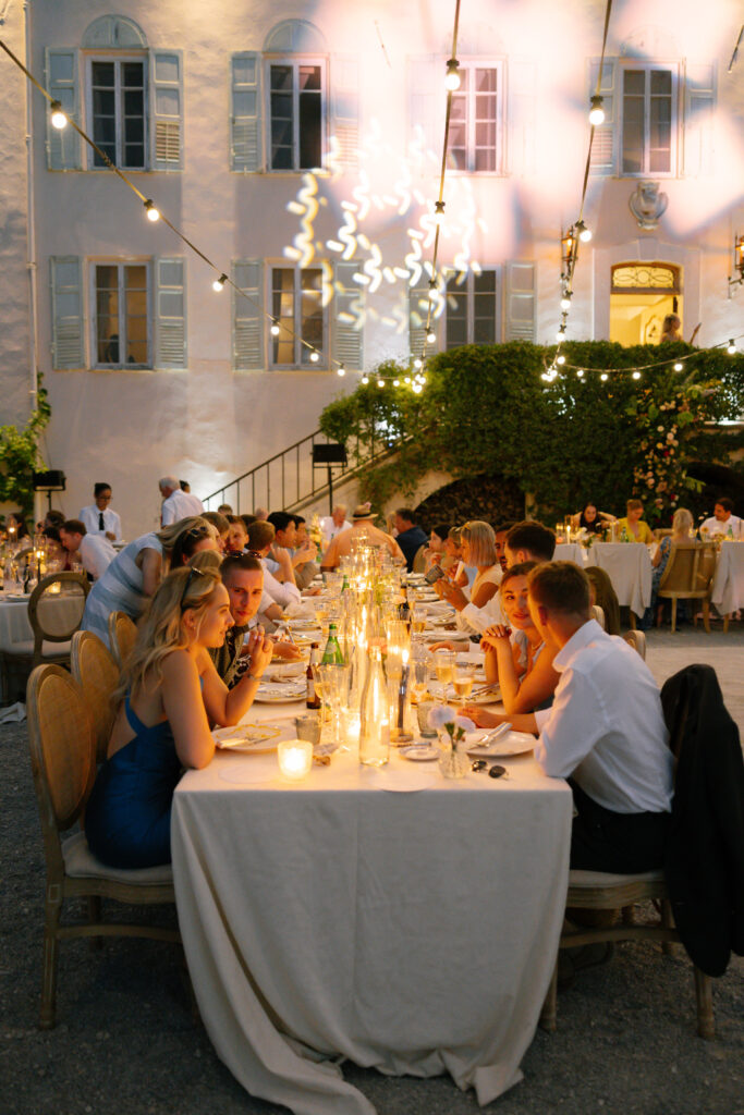 Guests sit at long, elegantly decorated tables with candles and string lights, dining outdoors in front of a large white building with shutters and greenery in the evening.
