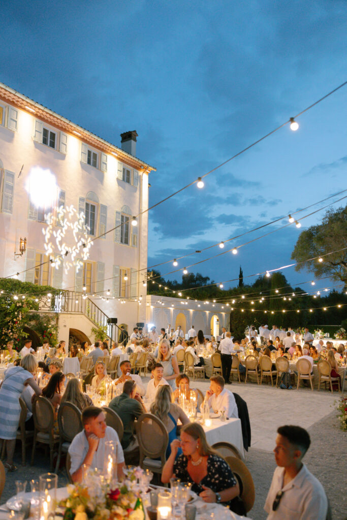 An outdoor evening event with guests dining at round tables under string lights beside a large white building. The atmosphere is festive and elegant, with candlelit tables and people engaged in conversation.