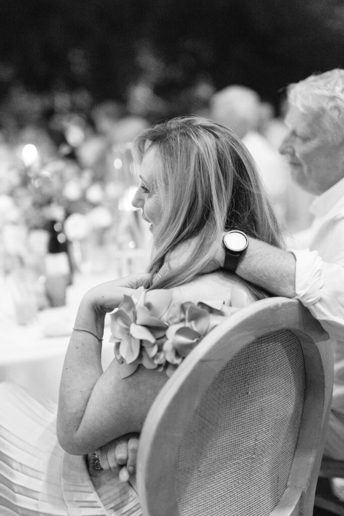 A woman with light hair sits at a table, smiling, with a mans arm around her shoulders. They are dressed formally, surrounded by blurred guests and decorations at what appears to be an event or celebration.