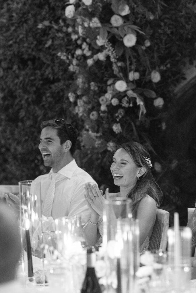 A man and a woman sit at a decorated table, smiling and clapping joyfully. The scene is elegant, with candles, flowers, and glassware, suggesting a celebratory event like a wedding or formal dinner.