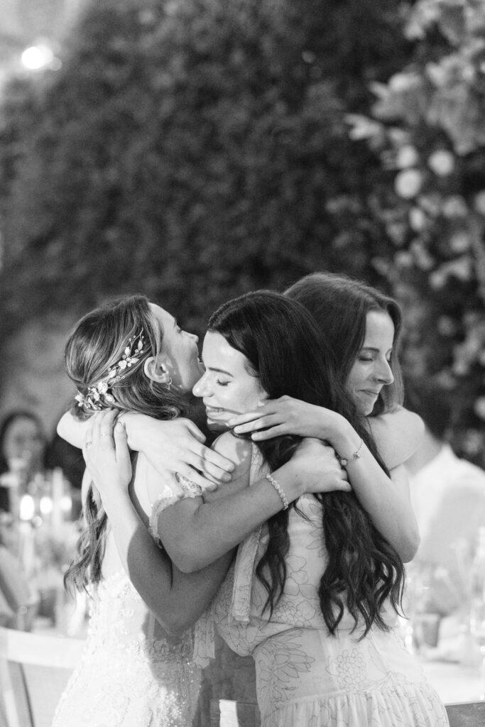 Three women in elegant dresses embrace in a joyful group hug at an outdoor event, likely a wedding. The scene is warmly lit with blurred greenery and candles in the background, creating an intimate atmosphere.