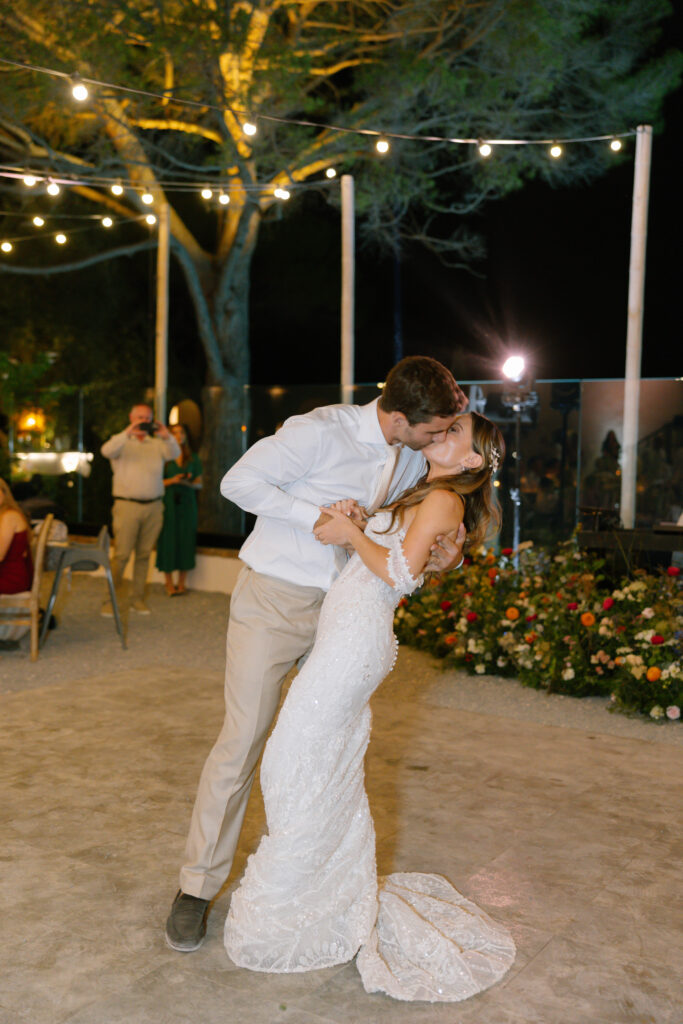A bride and groom share a kiss while dancing outdoors at night under string lights, surrounded by flowers and trees. The groom dips the bride as guests watch in the background.
