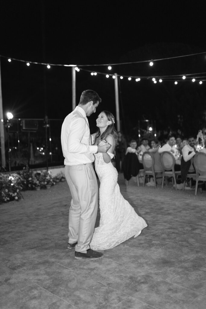 A bride and groom share a joyful first dance outdoors at night under string lights, surrounded by seated guests watching and smiling.