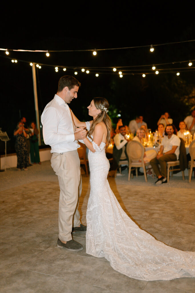 A bride and groom share their first dance outdoors at night under string lights, surrounded by seated guests watching and smiling. The bride wears a long white gown, and the groom is in a white shirt and tan pants.