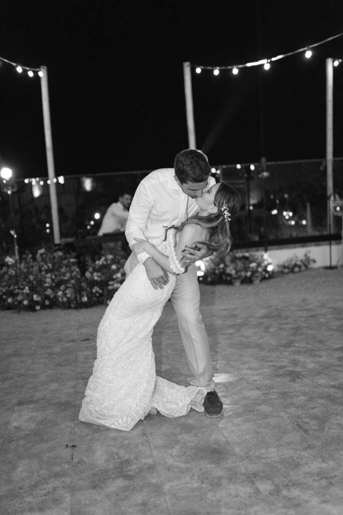 A groom in dress shirt and pants dips his bride, who wears a long lace gown, as they share a romantic kiss on a nighttime dance floor with string lights and flowers in the background.