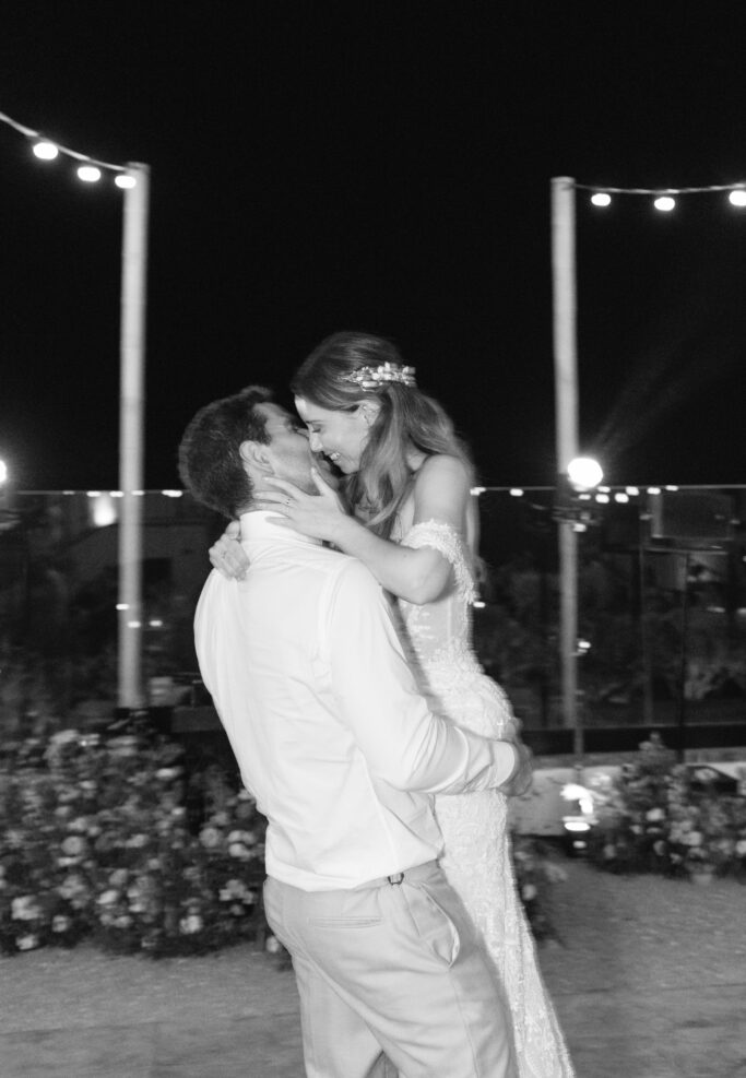 A groom lifts and embraces his bride while they smile and look into each others eyes during an outdoor nighttime wedding celebration, surrounded by string lights and floral decorations.