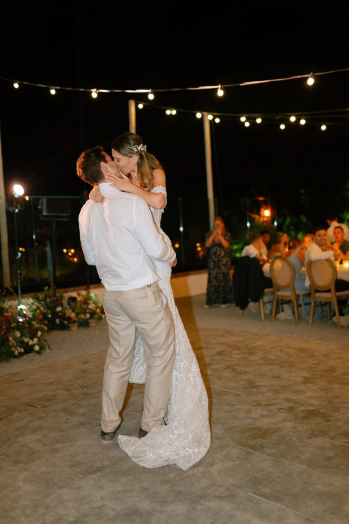 A bride and groom share a romantic first dance outdoors at night under string lights, surrounded by seated guests. The bride wears a white gown and the groom is in a white shirt and beige pants.
