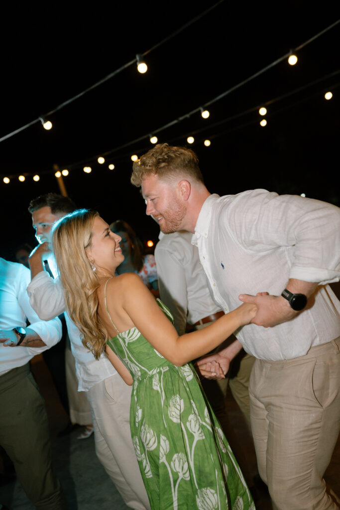 A smiling couple dances together outdoors at night under string lights, surrounded by other people. The woman wears a green floral dress and the man wears a white shirt and beige pants.