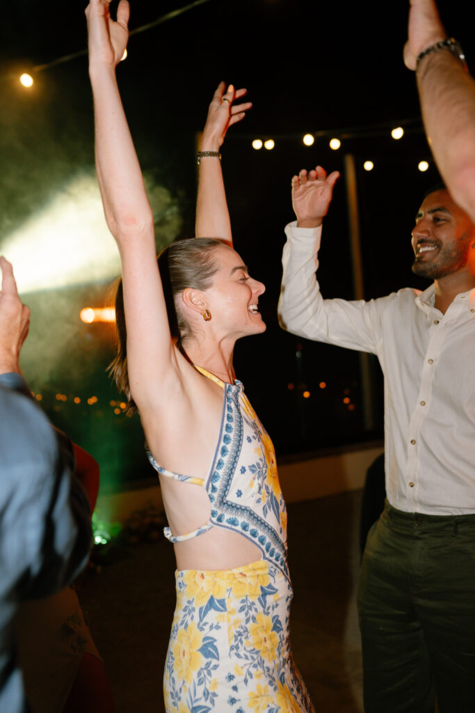 A woman in a floral dress dances with her arms raised, smiling, while a man in a white shirt dances beside her at an outdoor nighttime event with string lights in the background.