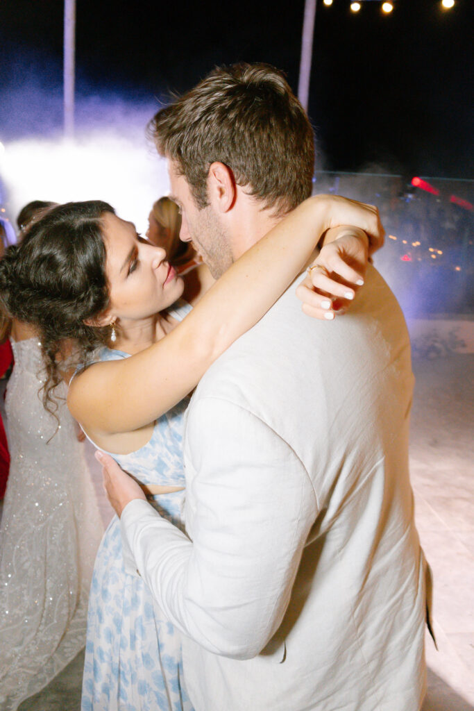 A woman in a light blue dress embraces and looks up at a man in a light-colored suit as they dance together at night, with soft lights and mist in the background.
