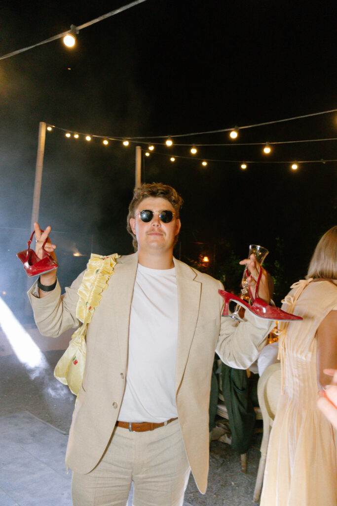 A man in a beige suit and sunglasses stands under string lights at night, holding red high heels and a drink, with a yellow bag on his shoulder. Theres a festive atmosphere with people and smoke in the background.