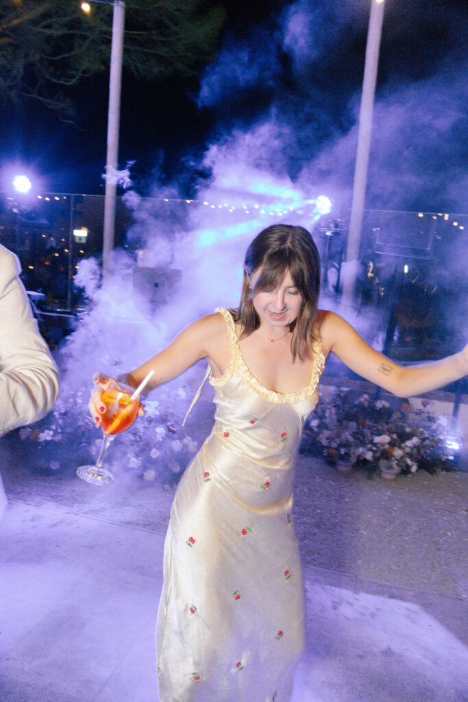 A woman in a light-colored, floral dress dances joyfully with a drink in hand, surrounded by smoke and colorful lights at an outdoor nighttime event.