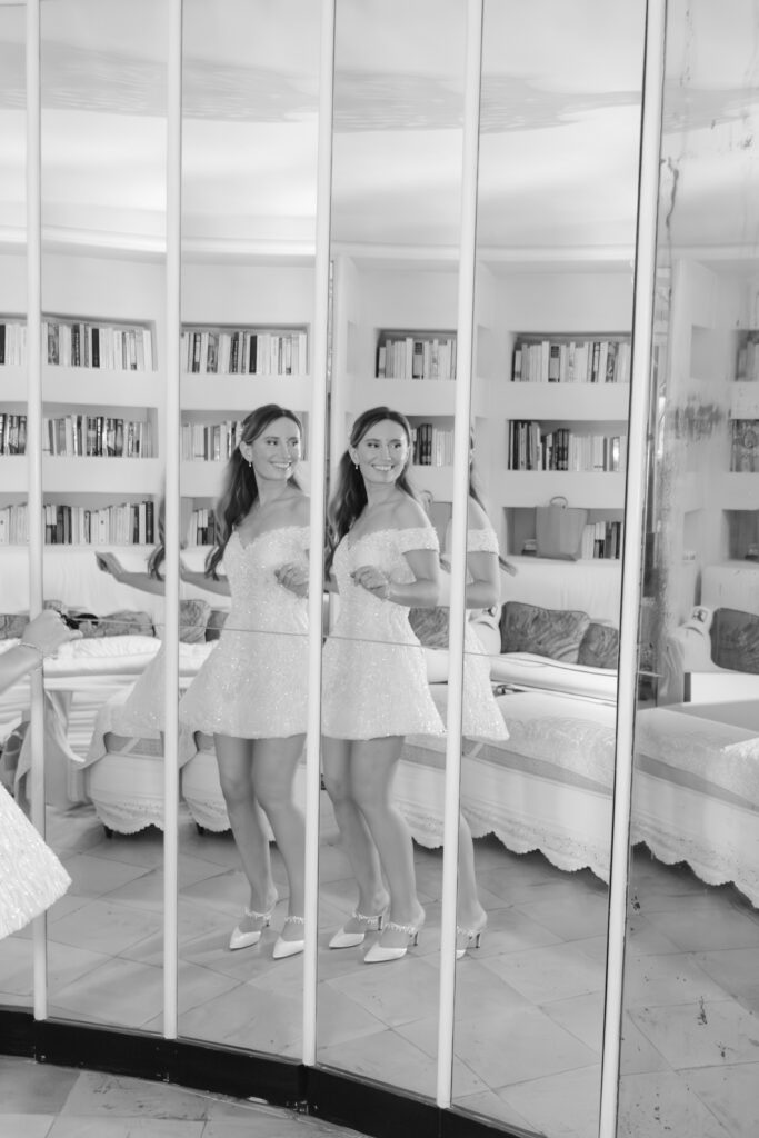 A woman in a short, white dress and heels stands in front of a set of mirrors, creating multiple reflections of her. The room has bookshelves, a sofa, and is decorated in a light, elegant style. The photo is in black and white.