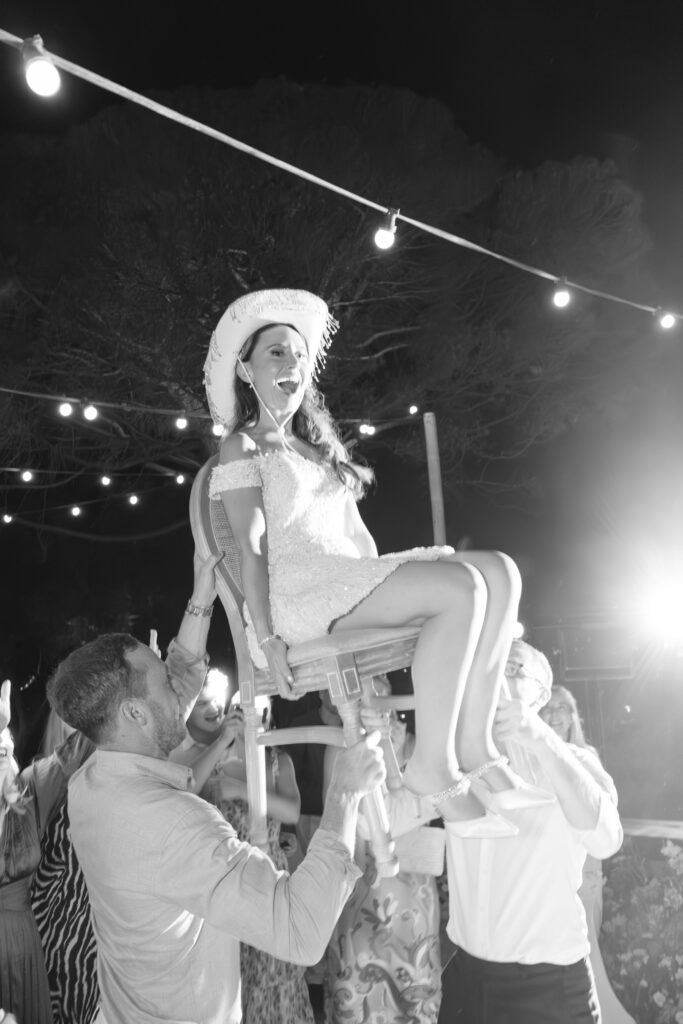 A joyful woman in a white dress and cowboy hat is lifted on a chair by guests during an outdoor celebration at night, with string lights illuminating the scene.
