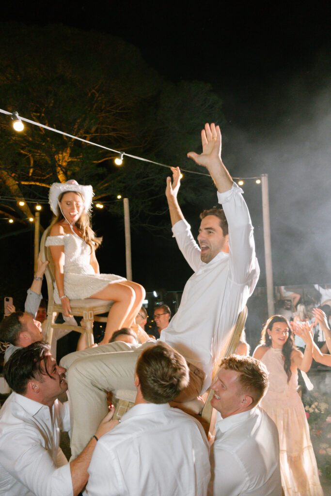 A bride and groom are lifted on chairs by guests during an outdoor nighttime celebration, surrounded by string lights and cheering friends. The bride smiles, wearing white with a playful hat, while the groom raises his arms joyfully.