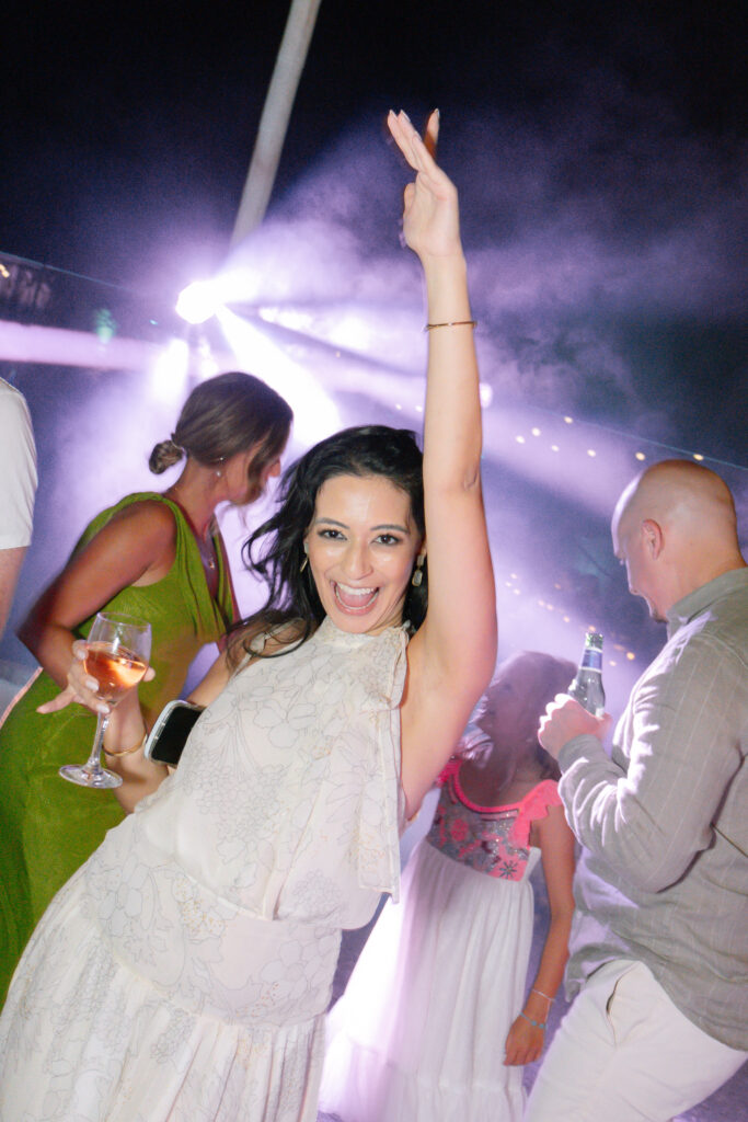 A woman in a white dress smiles and raises her arm excitedly while holding a drink at a lively outdoor party, with other people socializing and colorful lights in the background.