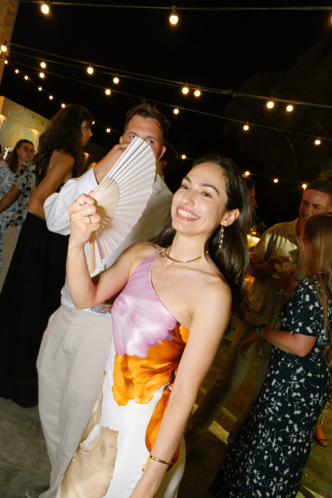 A smiling woman in a colorful dress holds a hand fan near her face at an outdoor evening event, surrounded by people under string lights.