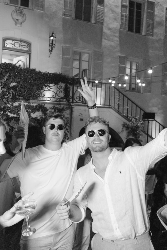 Two men wearing sunglasses and light-colored shirts smile and pose for a photo at a lively outdoor party at night, holding drinks and fans, with string lights and a staircase in the background.