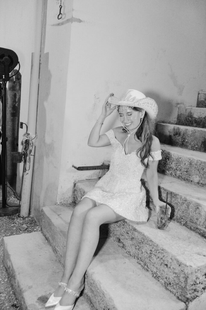 A woman in a short, off-shoulder white dress and cowboy hat sits on stone steps, smiling and holding a drink. The scene is in black and white, and she touches the brim of her hat with one hand.