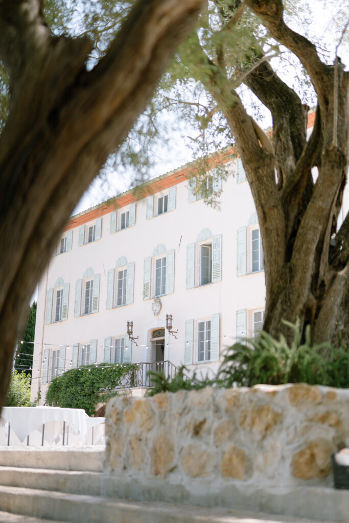 A large Mediterranean-style building with light blue shutters and a terracotta roof is seen through the branches of tall trees, with stone steps and a low stone wall in the foreground. Bastide Du Roy