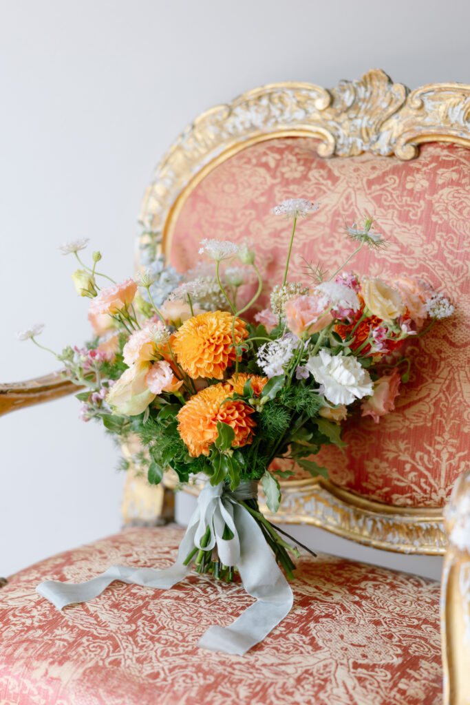 A vibrant bouquet of orange, white, and pink flowers with greenery rests on an ornate gold and red upholstered chair, tied with a pale blue ribbon.