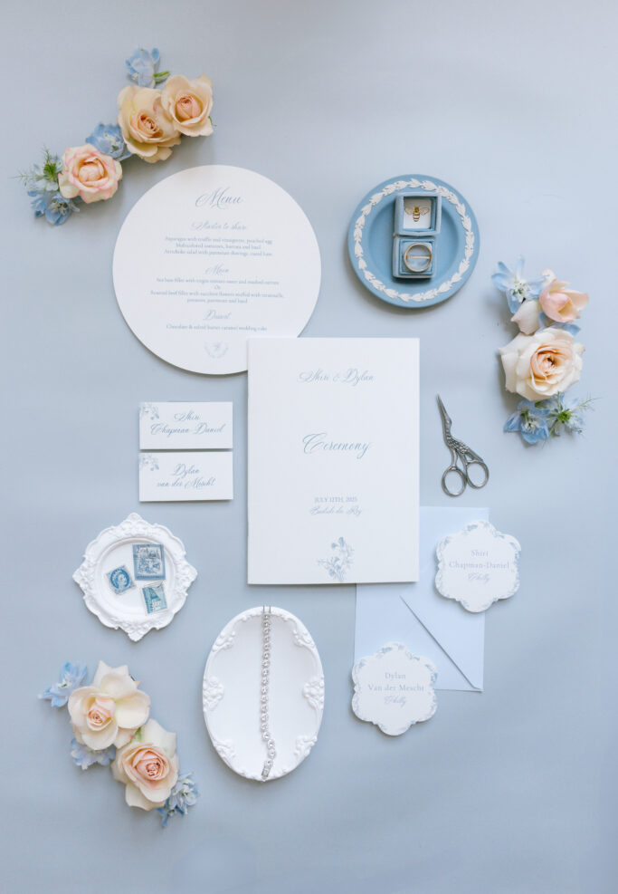 Elegant wedding flat lay with invitations, menu, RSVP cards, jewelry, scissors, a ring box with rings, and pastel flowers arranged on a pale blue background.