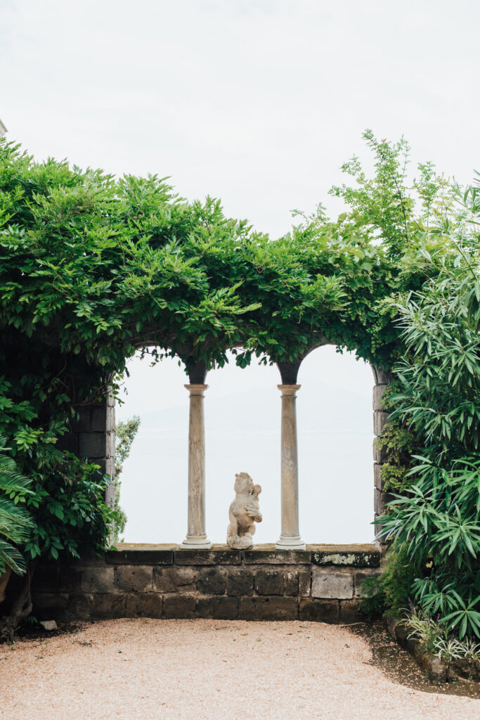 A stone terrace with two columns and lush greenery surrounds a small statue of a cherub, overlooking a bright, hazy body of water in the background.