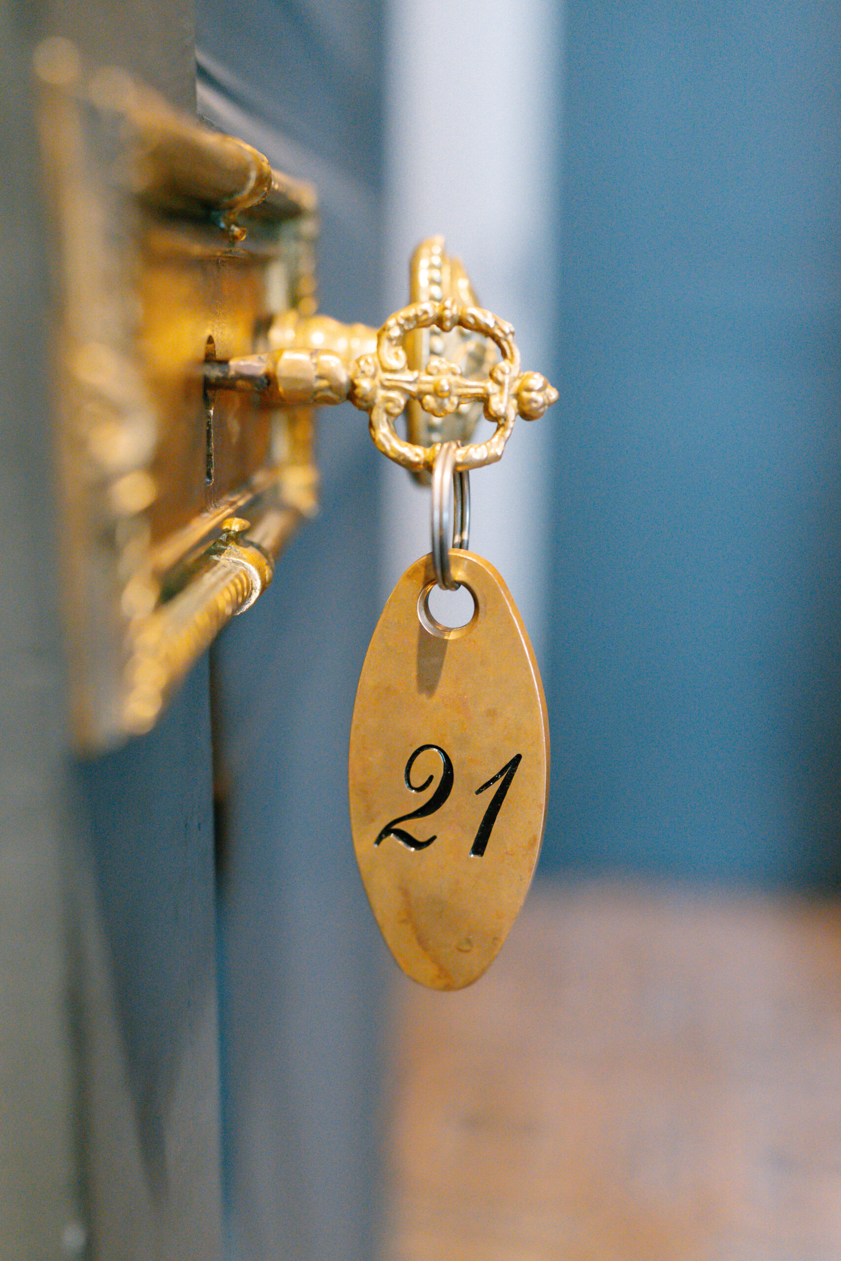 A vintage gold key with an ornate design is inserted into a lock, with a brass key tag labeled 21 hanging from it. The background is slightly blurred in blue and brown tones.