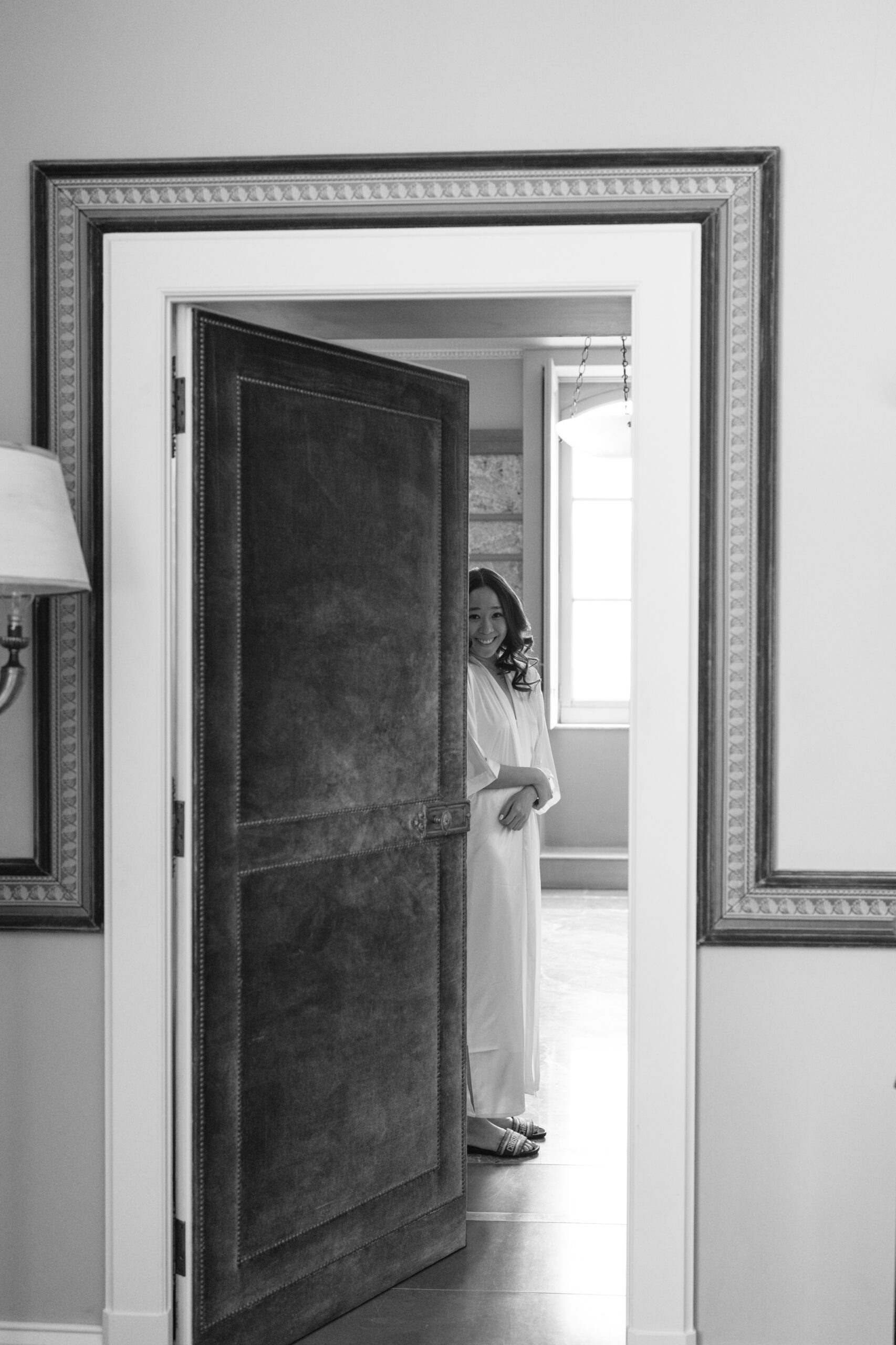 A woman in a white robe stands in a bright room, partially visible through a slightly open dark door, smiling softly and looking toward the camera. The scene is in black and white.