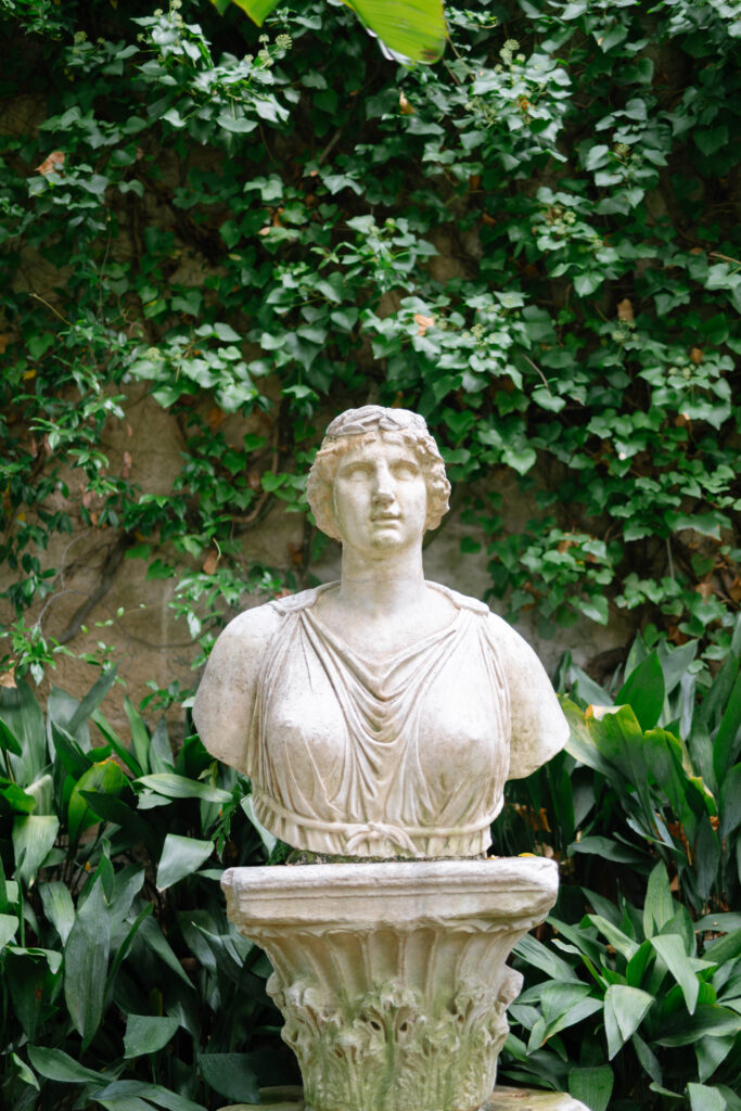 A marble bust of a woman wearing draped clothing stands on a pedestal surrounded by lush green plants and ivy-covered walls.
