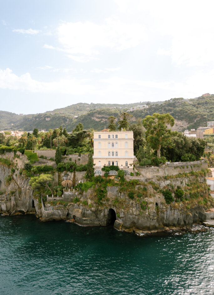 A large, pale yellow villa with green shutters sits atop rocky cliffs surrounded by lush greenery, overlooking clear blue water, with hills in the background under a partly cloudy sky.