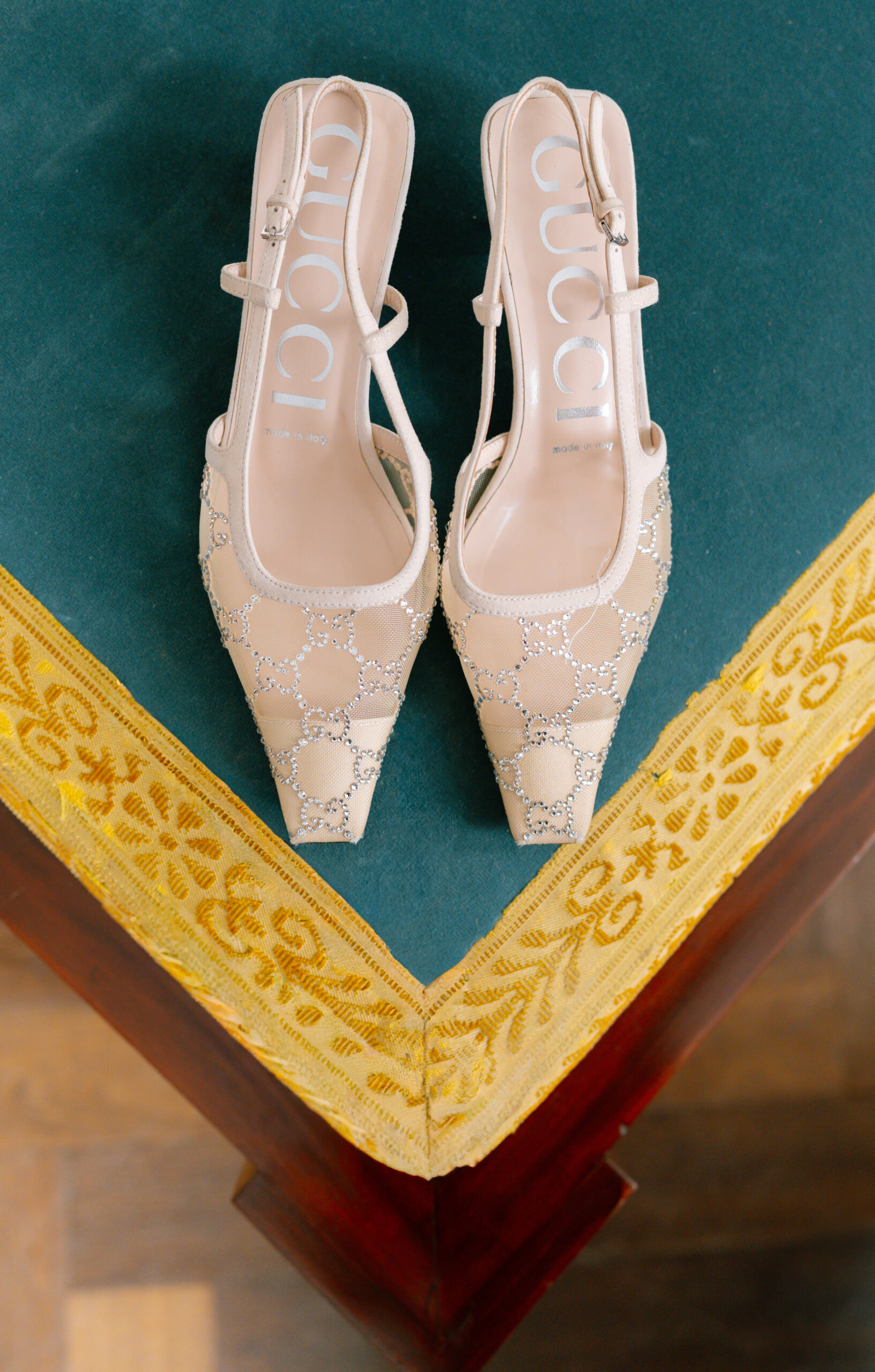 A pair of elegant, cream-colored Gucci heels with sparkling embellishments are displayed on a green velvet surface with a yellow embroidered border.