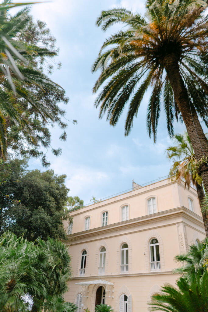 A beige, three-story building with arched windows and balconies is surrounded by tall palm trees and lush greenery under a partly cloudy sky.