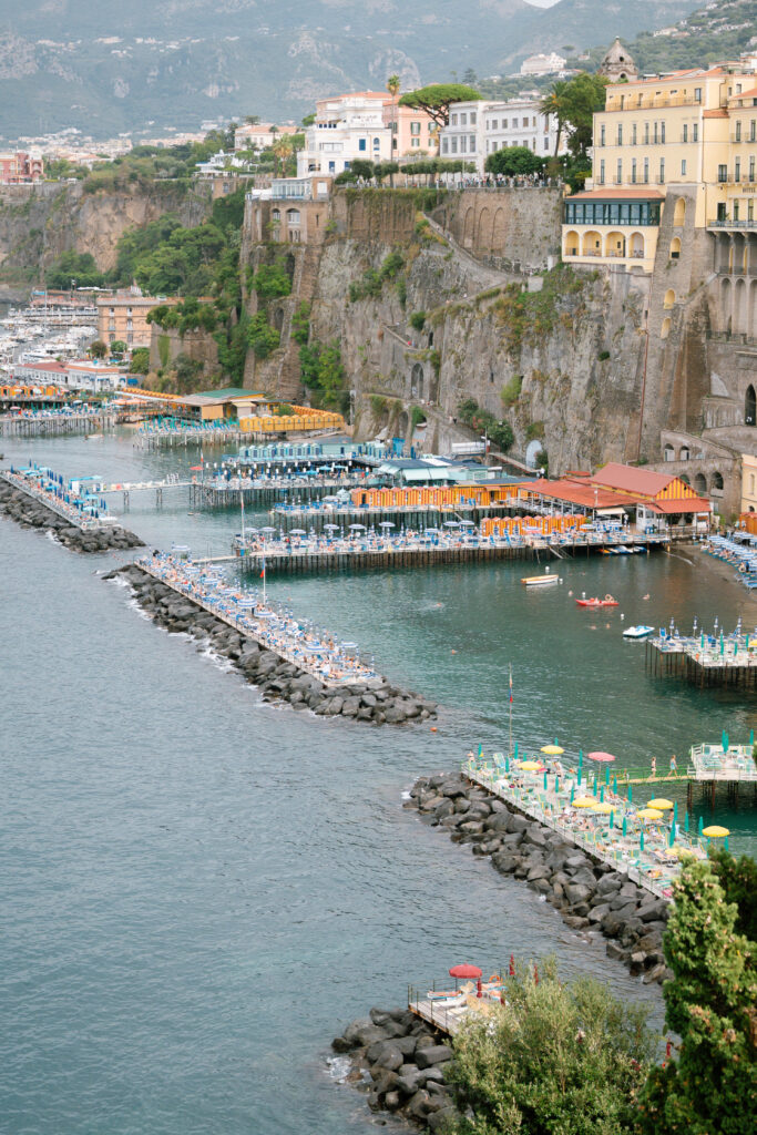 A coastal view of Sorrento, Italy, featuring colorful beach umbrellas and sunbathers on piers extending into the blue water, with cliffs and historic buildings rising above the shoreline.