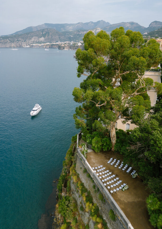 Rows of white chairs set up for an outdoor ceremony on a cliffside terrace overlooking blue water, with a large tree, lush greenery, a white boat in the sea, and mountains in the distance.