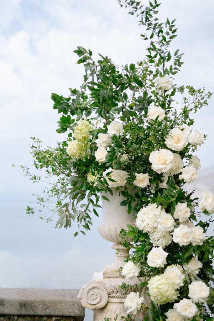 A large white urn overflowing with white roses, hydrangeas, and greenery sits outdoors against a cloudy sky, creating an elegant, lush floral arrangement.