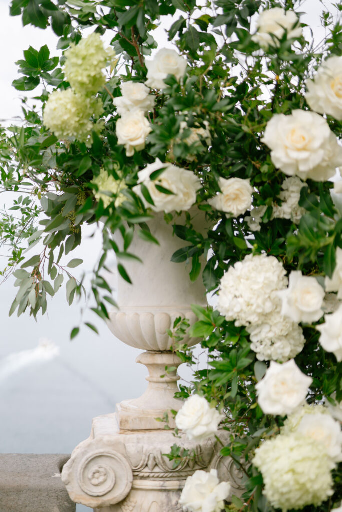 A large, ornate stone vase filled with lush white roses, hydrangeas, and green foliage, set outdoors with a blurred background, possibly near water.